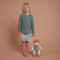 A curly, blonde haired child stood next to an Olli Ella Pea Dinkum Doll which is also standing up. The child is pointing at the doll