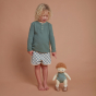 A curly, blonde haired child stood next to an Olli Ella Pea Dinkum Doll which is also standing up. The child is pointing at the doll