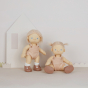 Two Olli Ella Petal Dinkum Doll pictured against a light pastel coloured background. One doll is stood up, the other sat down, wearing their pink hair in bunches with cut out house and grass shapes behind them 