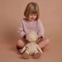 A child sat down cross legged with a Olli Ella petal Dinkum Doll sat in front of them. The child is playing with the dolls hair 