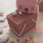 Olli Ella Medium Piki Basket in natural rattan, a rattan picnic basket with white handles, sat on a pastel checked picnic blanket with the an Olli Ella Case Rose bag and 2 wooden eggs sat on top, sun shining through in the background.
