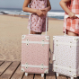 A child on a beach holding an Olli Ella Pink Daisies See-Ya Suitcase with another child next to them holding the leafed mushroom print one 
