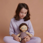 A child sitting cross legged holding the Olli Ella Bitsy Dinkum Doll on their lap. The child is looking down at the doll. Both child and doll are wearing purple clothing. Pictured on a plain terracotta coloured background.