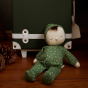 Olli Ella Dozy Dinkum Pudding in a pale green outfit, with leaf pattern sat in front of a dark green suitcase with white detail