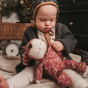Olli Ella Dozy Dinkum Limited Edition Pudding, held by a baby wearing a rib knit grey cardigan and patterned bonnet, an Olli Ella wagon in the background with a Christmas tree. All sat on a natural stripy muslin