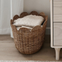 The Olli Ella Rattan Tulip Storage Basket in Natural, on a rug in a bedroom, holding a blankets.