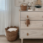 The Olli Ella Rattan Tulip Storage Basket in Natural, on the floor next to a chest of draws, holding blankets.