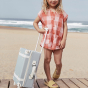 Child standing on decking on a sandy beach holding the Olli Ella Steel Blue See-Ya Suitcase
