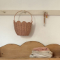 The Olli Ella Rattan Tulip Carry Basket - Seashell Pink hanging on a coat rack above a wooden shelf with folded blankets