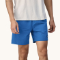 A front, close up of an adult wearing the Patagonia Men's Baggies Lights - Endless Blue. Mid-Blue baggy men's shorts, and a white t-shirt on a cream background