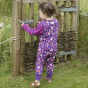 Young girl stood next to a wooden gate wearing the pink and purple Piccalilly dungarees in the woodland treasures print