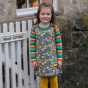 a girl wearing organic cotton pinafore dress with a fun rainbow weather and planets all-over print and a rainbow-striped long sleeve top from piccalilly