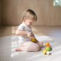 Toddler cutting a play food lemon with a wooden knife, kneeling on a rug.