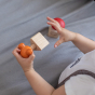 Child holding Orange Tap Bolt of the PlanToys Wooden Nuts and Bolts puzzle in a wooden Triangle block. The Plan Toys Red Button Bolt is below in a wooden square block