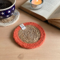 Close up of an orange jute and seagrass Respiin coaster on a wooden table next to an open book