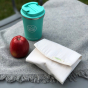 A Slice of Green reusable sandwich wrap on a grey blanket next to an apple and a reusable coffee cup