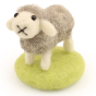 A fully made The Makerss - Amiguwoolli Tiny Chicken Mini Needle Felt Figure. The sheep is made from grey woolen felt, with a cream head, ears and legs, stick in eyes and a black mouth, and is stood on  alight green felted base.