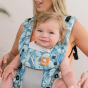 Tula - A woman carries a child on her chest wearing a Coast Paradise - Explore Baby Carrier. This is a front-on lifestyle shot with a soft beige background. The focus is on the button detail on the carrier.