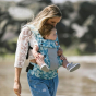 Tula - A woman carries a child on her chest wearing a Coast Paradise - Explore Baby Carrier. This is a side-on lifestyle shot in an outdoor environment. The baby is facing forward in the carrier.