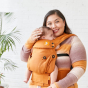 A female carrying a baby in the Tula Hemp Explore baby carrier, front inwards facing, female in multitude orange and rose top, hand on babies head and cheek touching, white brick wall and large plant behind. 