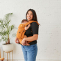 A female carrying a baby in the Tula Hemp Explore baby carrier, front inwards facing, female in black top and jeans, white brick wall and large plant behind. 