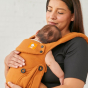 A female carrying a baby in the Tula Hemp Explore baby carrier, front inwards facing, female in black top, hand on baby, neutral wall behind. 