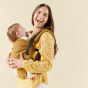 A female carrying a baby in the Tula Hemp Free to Grow Olivine baby carrier, front inwards facing, female in yellow patterned top and white jeans, laughing, cream wall behind. 
