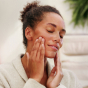 A person applying the Weleda Skin Food Nourishing Cleansing Balm to their face, their eyes are closed 