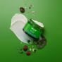 An open pot of Weleda Skin Food Nourishing Night Face Cream, the product can be seen spilling out the top. The top is placed on a green surface surrounded by ingredients 
