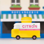 Picture of the Citron Candylab Candyvan with a high street store in the background. It is a 1960s style yellow and white van with a yellow Lemon Macaron on the roof.