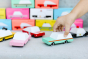 Close up of a childs hand holding the Candylab handmade teal wagon toy vehicle next to the pink sedan and taxi toys