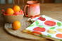 Close up of the DUNS Sweden organic cotton linen tea towel in the citrus print on a wooden table next to a bowl of toy oranges and a red mug