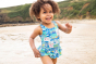 Child running around on a beach wearing the Frugi Postcards design Little Coral Swimsuit