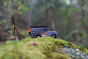 Candylab Drifter Kodiak, a 4x4 off-roader wooden car toy, in grey and black, with metal roof rack and rubber tyres, on mossy, rocky ground with out of focus trees in background, side view