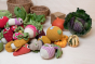 Myum handmade soft crochet vegetable toys scattered in a pile on a white table next to come real vegetables and wooden baskets