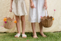 Two children wearing sandals and dresses stood on the grass holding Olli Ella Rattan Tulip Carry Basket - Seashell Pink and Natural filled with flowers from the garden