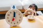 Young child crouched down holding a teddy bear next to an empty bowl beside the PlanToys wooden activity clock