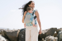 Woman stood in front of some rocks wearing the Tula blue and beige Coast Paradise FTG baby carrier