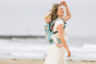 Woman walking along a beach wearing a baby on her back in the Tula soft structured FTG Coast Paradise baby carrier