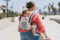 Woman walking on a beach wearing a baby on her back in the Tula FTG Coast Isle soft structured baby carrier