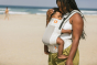 Close up of woman on a beach wearing the Tula beige Coast Isle FTG baby carrier