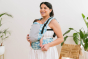 Woman stood in a white room wearing the Tula Coast Paradise soft structured baby carrier