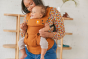 A female carrying a baby in the Tula Hemp Explore baby carrier, facing front, outwards facing, female in floral multicolour top and jeans, looking at baby beige wall behind. 