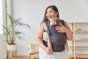 Woman stood holding a baby in a Tula organic cotton FTG baby carrier, in a dark grey colour
