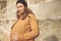 Mamalila Eco Wool Oslo Babywearing Coat in Camel. A light tan boiled wool winter babywearing coat. Side lifestyle view, with pregnancy insert. Stone wall background.  