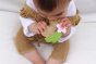 Close up of a toddler holding and chewing on the oli and carol aly the almond teething toy on a white blanket