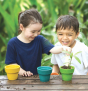 Two children add soil to the PlanToys Flower Pot Set and a plant. They are outside with a lush green background.