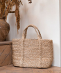 Turtle Bags Jute Basket stood upright against a cream wall, on a wooden floor and next to a wooden step with a clay pot containing dried twigs, leaves and catkins