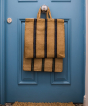 Turtle Bags Blue and Black Stripey Bag. A natural jute bag with short handles and 3 vertical blue and black stripes. Hanging off a central handle on a royal blue door with jute doormat on the floor