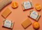 Bio-D natural Vegan mandarin soap bar on a light orange background, surrounded by mandarin oranges, soap bar boxes and a wooden brush